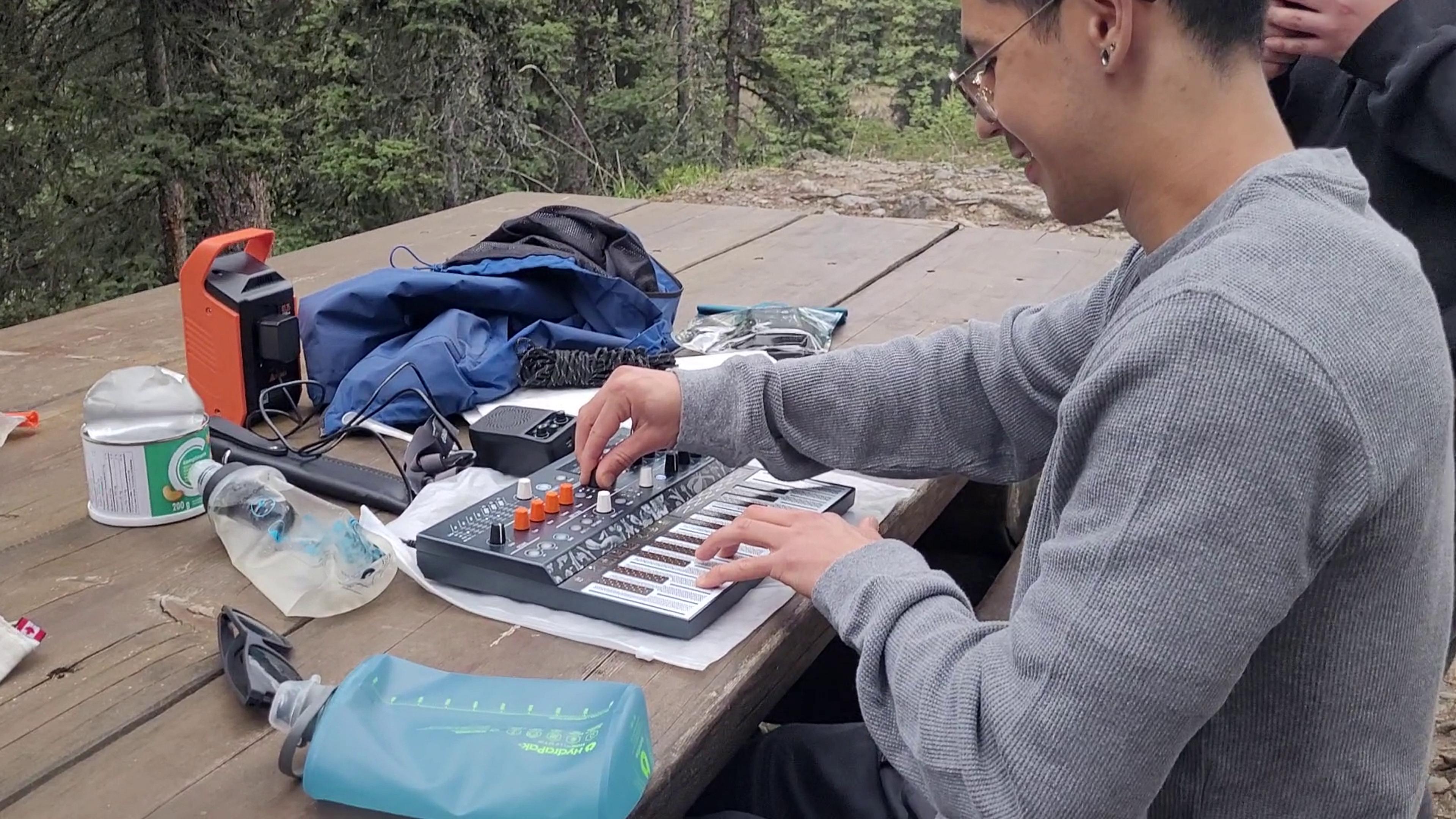 A person plays a synthesizer on a picnic table surrounded by forest scenery, possibly in a park or nature reserve.