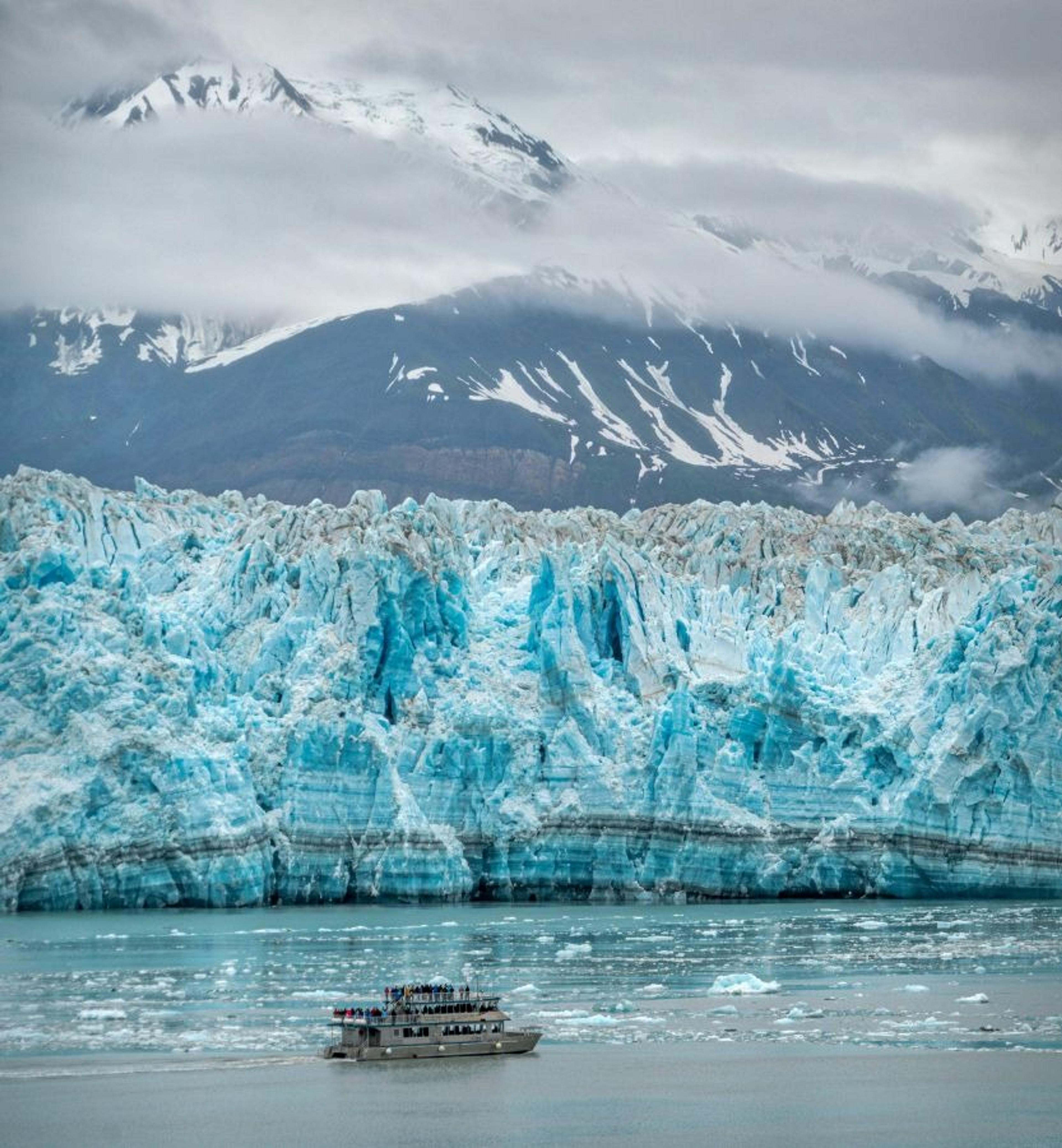 A tour boat navigates the icy waters in front of Alaska's Hubbard Glacier, with snow-capped mountains shrouded in clouds in the background.