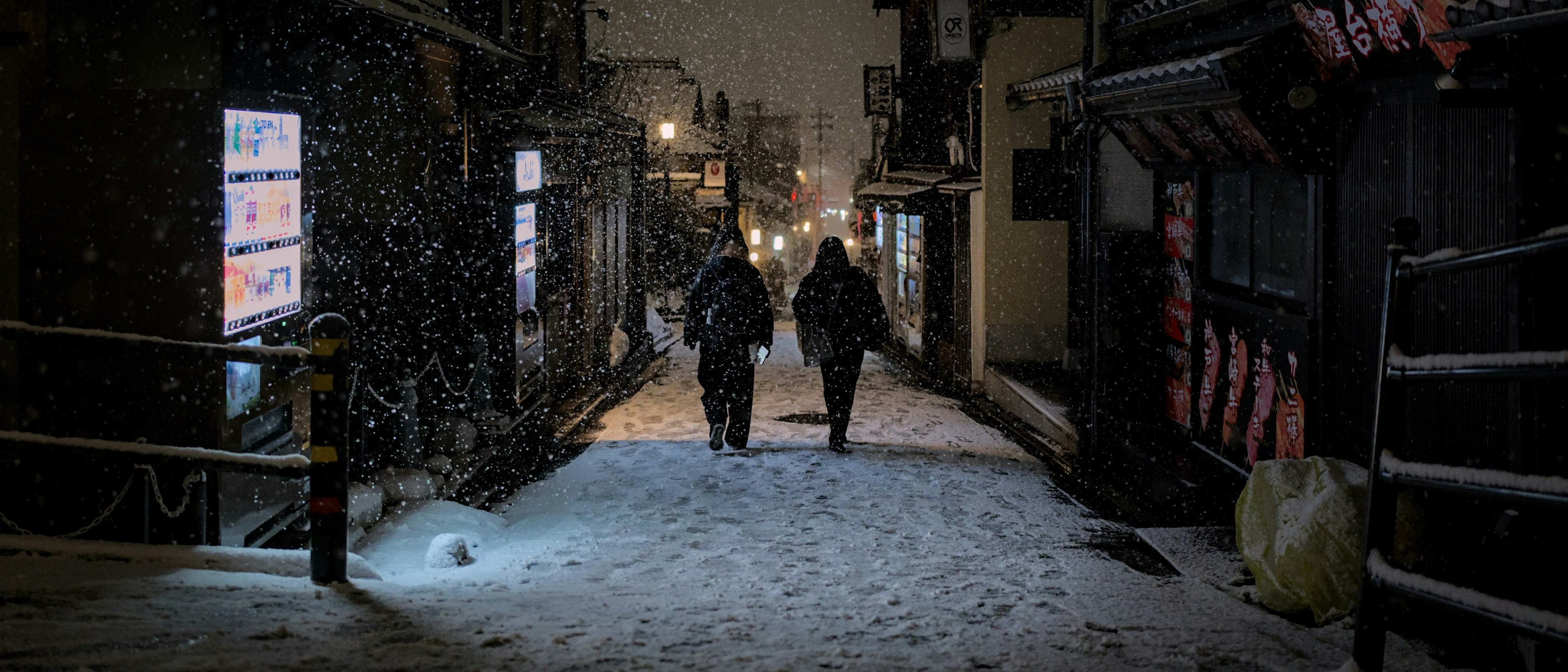 A snowy night scene unfolds on a narrow street in Kyoto, Japan, with illuminated signs and two people walking amidst the falling snow.