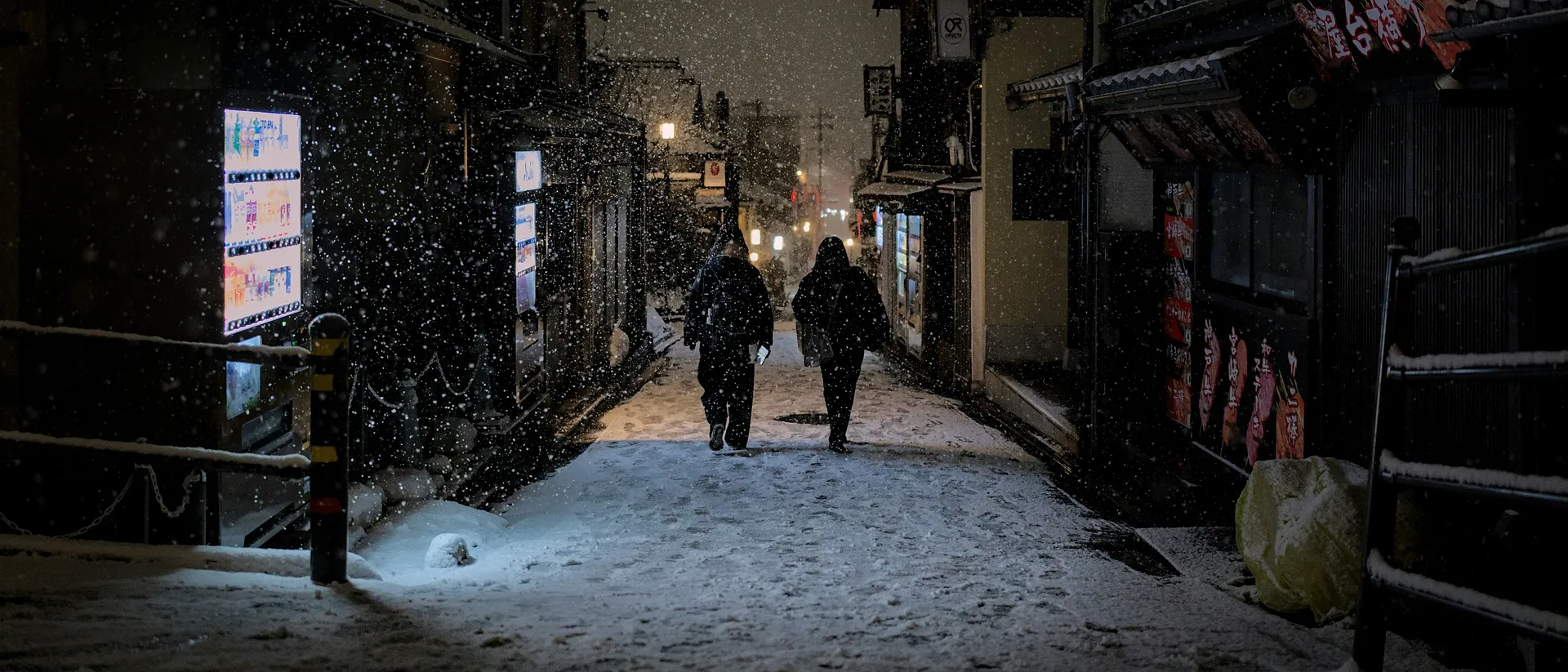 A snowy night scene unfolds on a narrow street in Kyoto, Japan, with illuminated signs and two people walking amidst the falling snow.