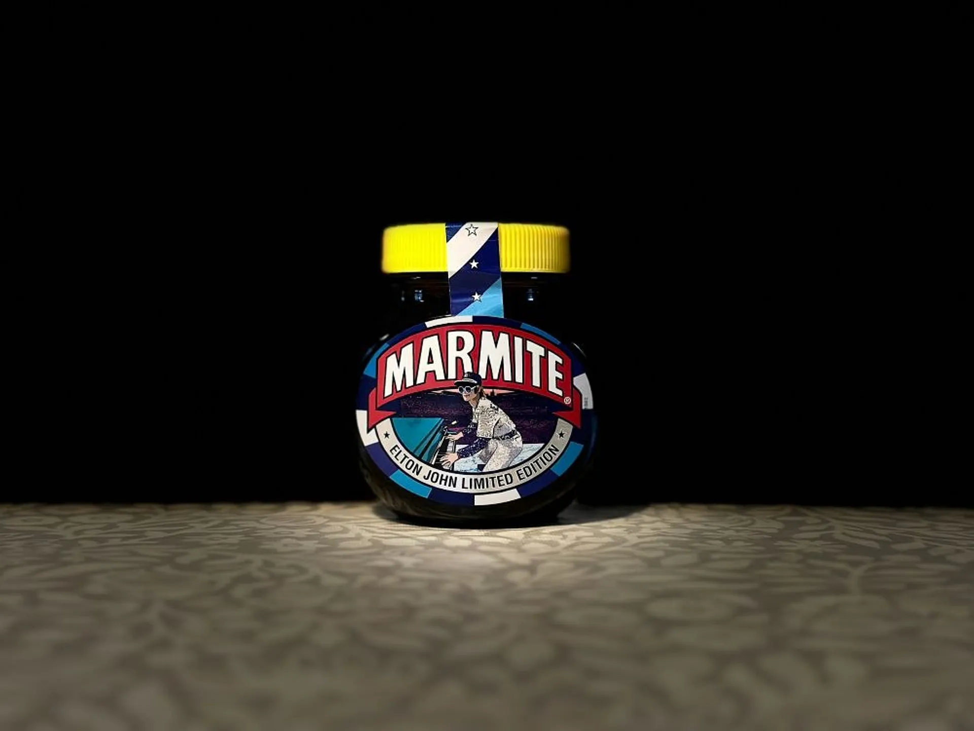 A limited edition jar of Marmite featuring a design inspired by Elton John stands on a patterned surface against a dark background.