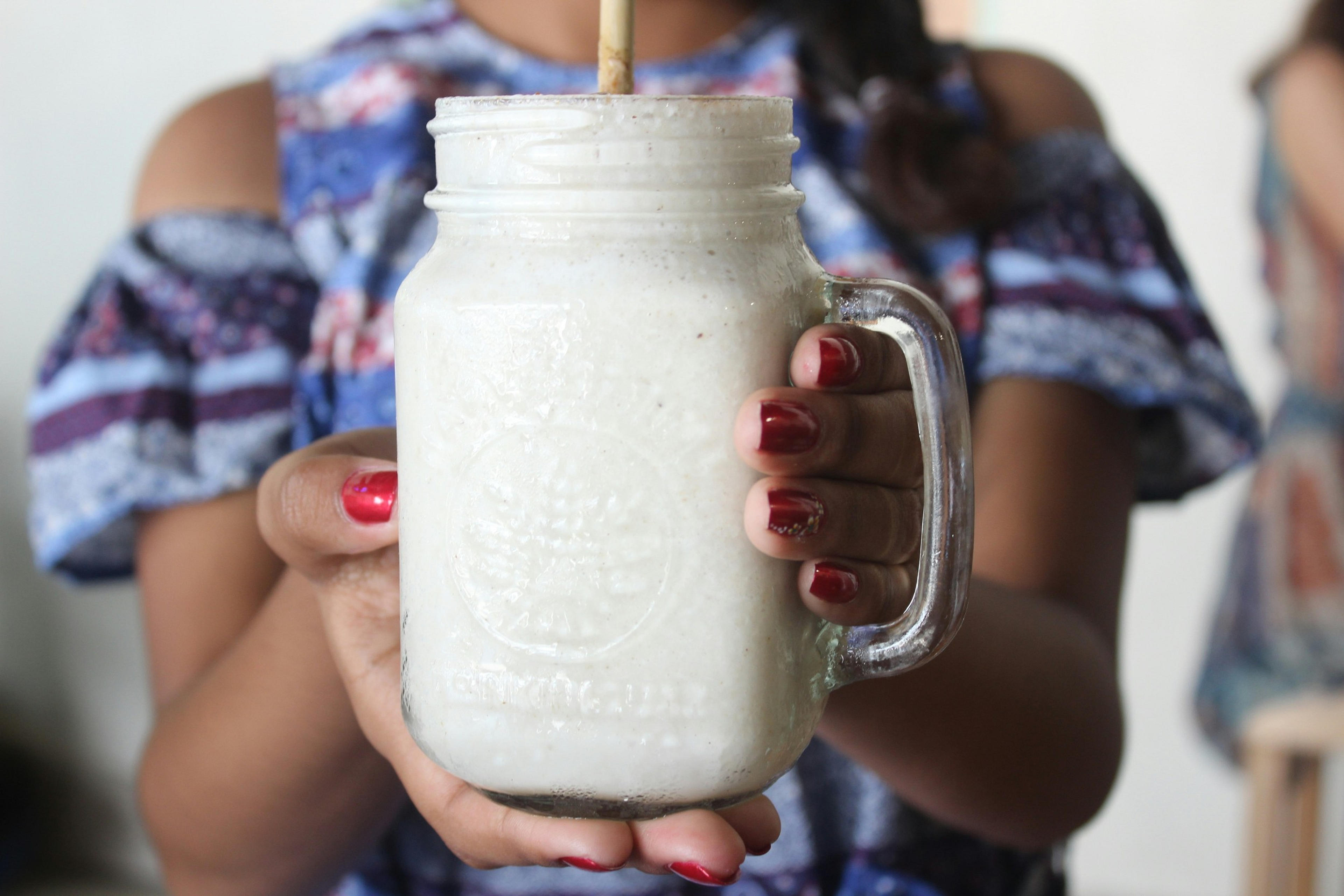 A close-up captures a person offering a creamy milkshake in a glass jar, with a focus on their red-painted nails.