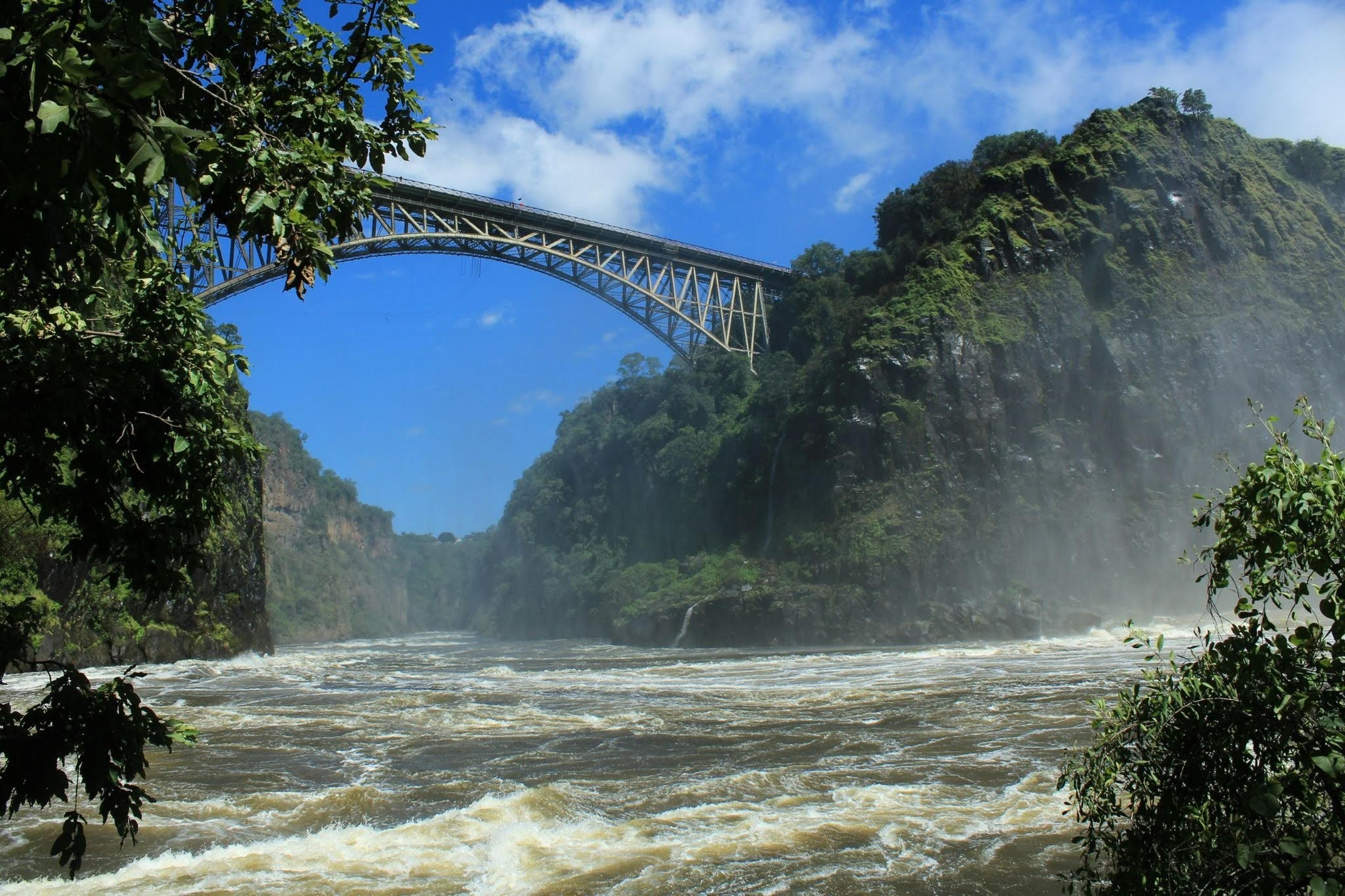 A steel arch bridge spans a turbulent river gorge near Victoria Falls, bordered by lush green cliffs under a clear blue sky.