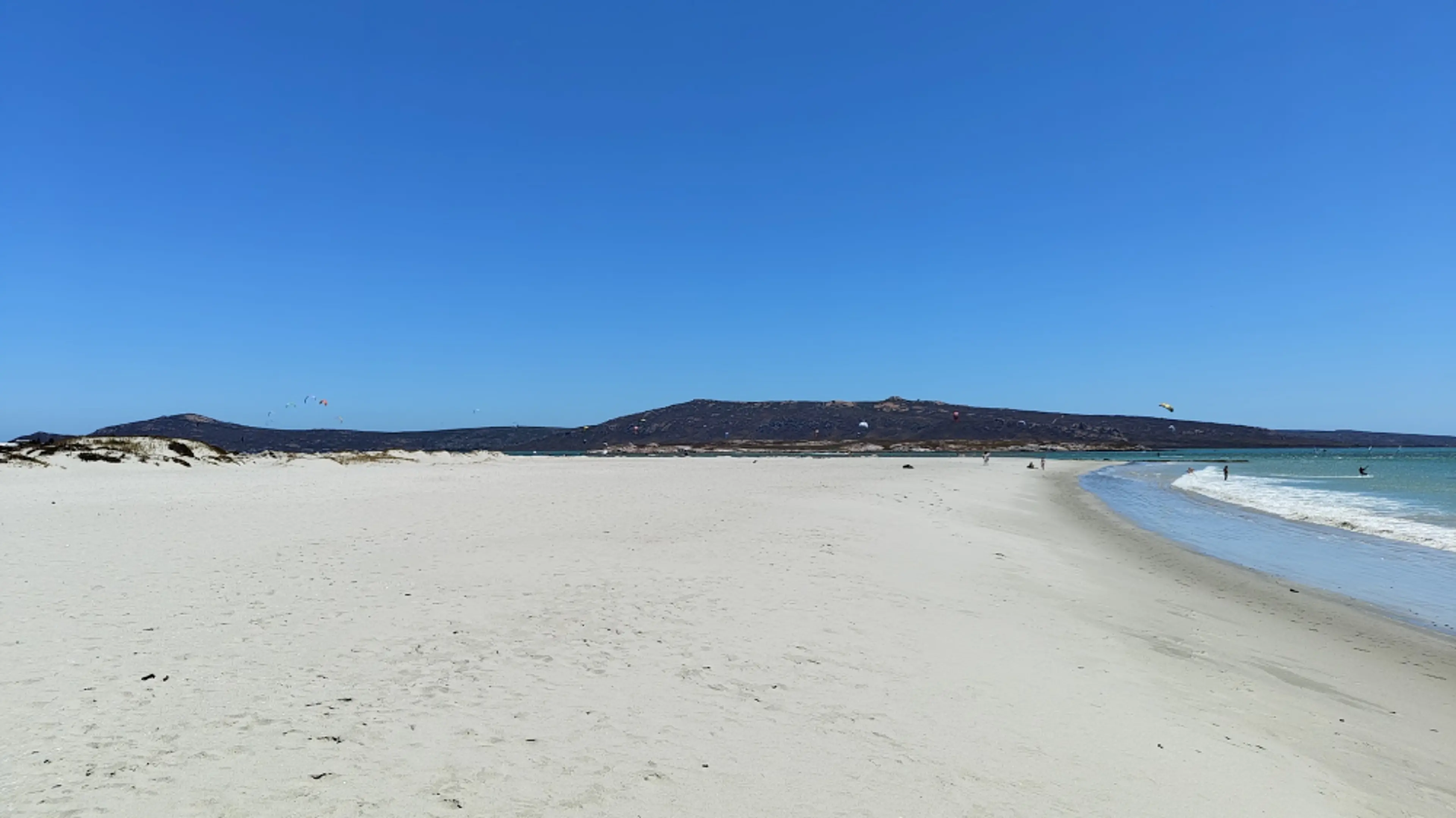A pristine sandy beach with kite surfers in Langebann, South Africa, stretches under a clear blue sky.