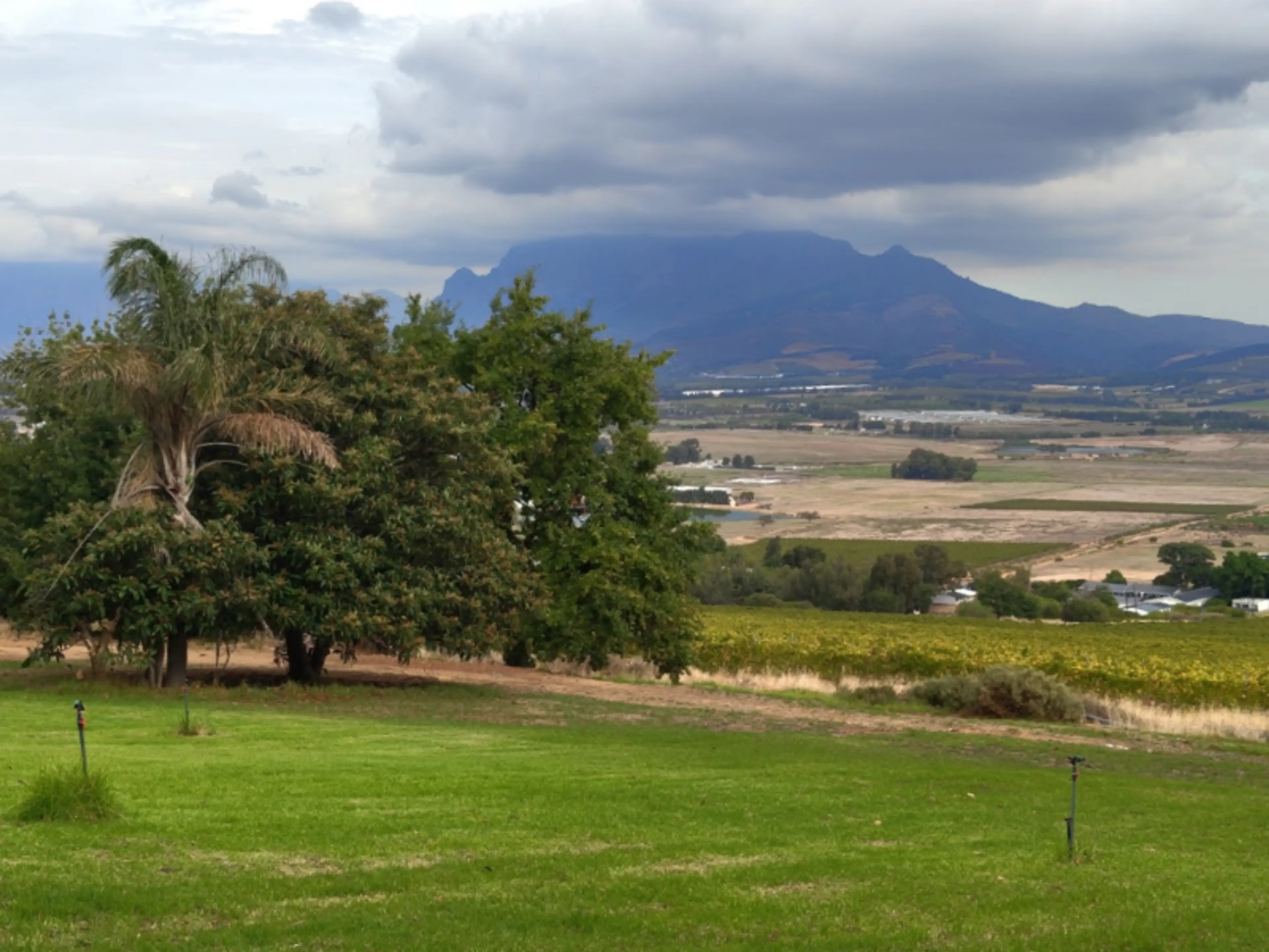 A lush green lawn and trees frame a view of the scenic Paarl valley in South Africa, with mountains in the background beneath a cloudy sky.