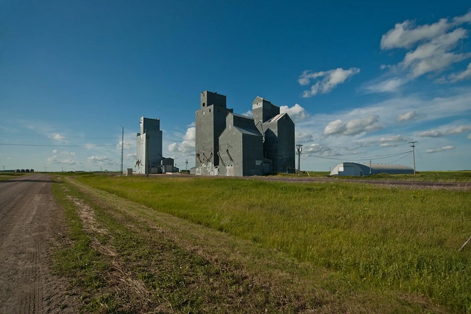 A vast prairie landscape in Saskatchewan features towering, weathered grain elevators under a bright blue sky with scattered clouds.