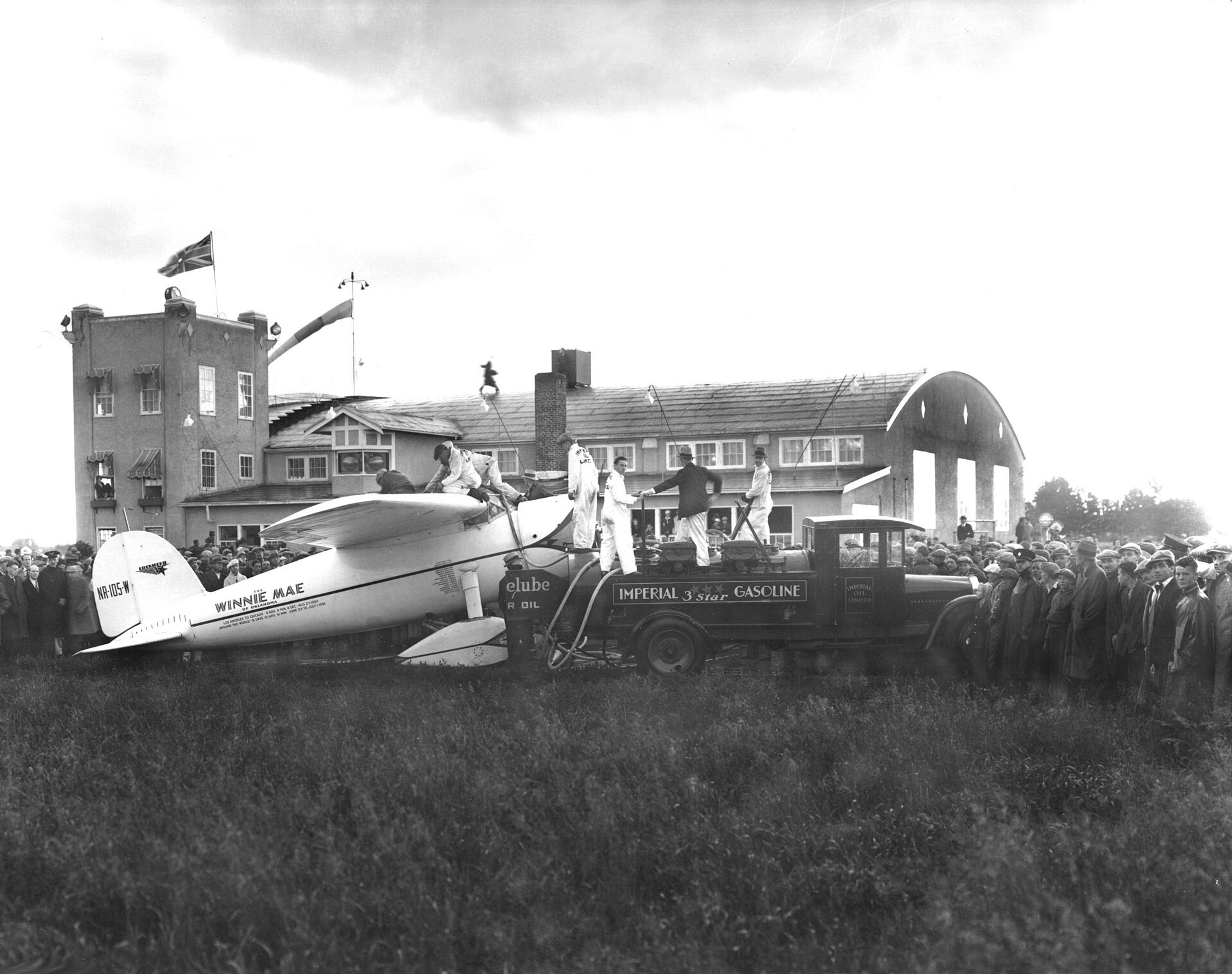 A fuel truck refuels the "Winnie Mae" monoplane at Edmonton Municipal Airport, Canada, surrounded by a crowd of onlookers.