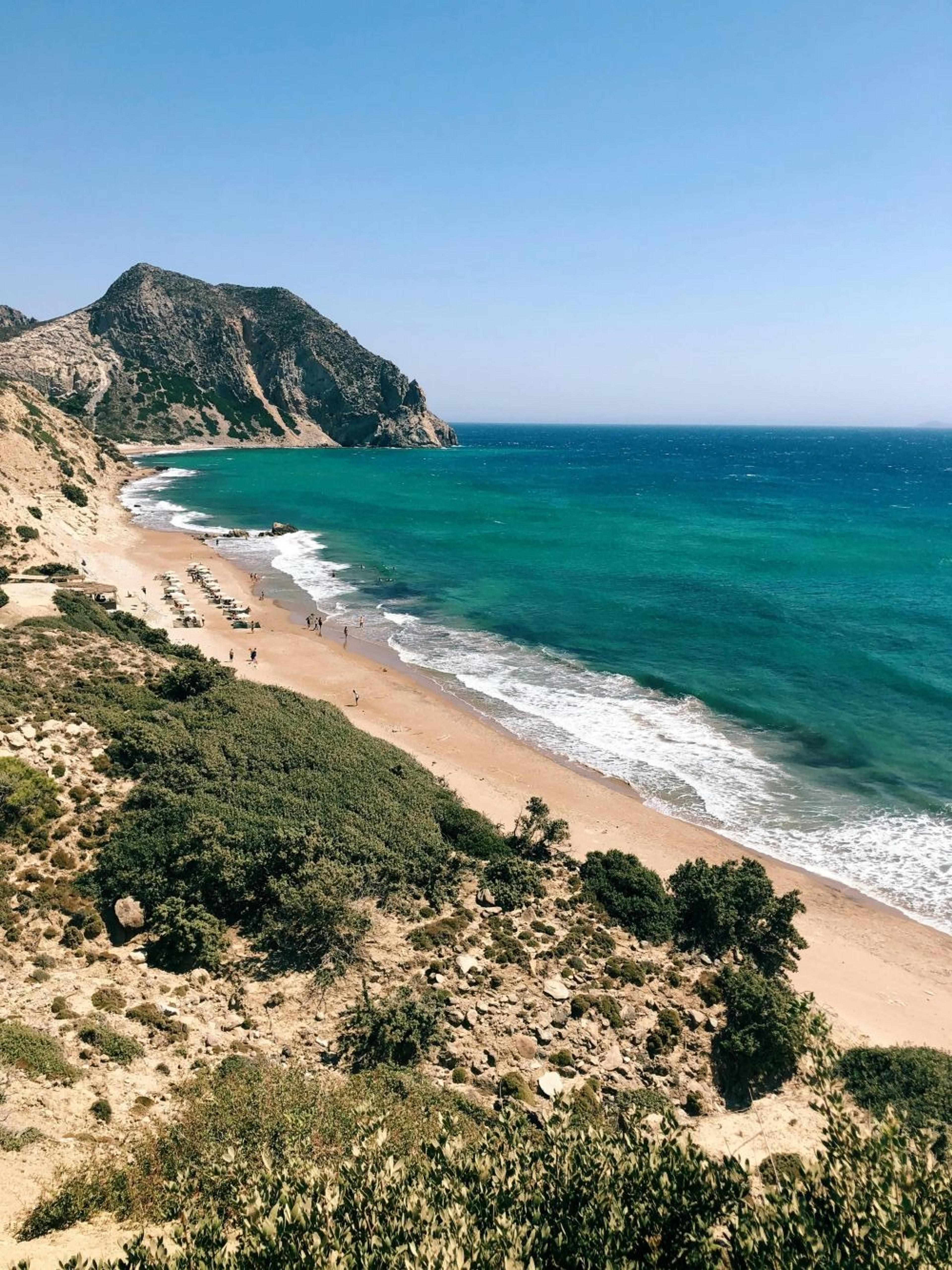 Turquoise waves lap against the sandy shore of Paradise Beach in Kos, Greece, framed by rugged hills and sparse greenery.