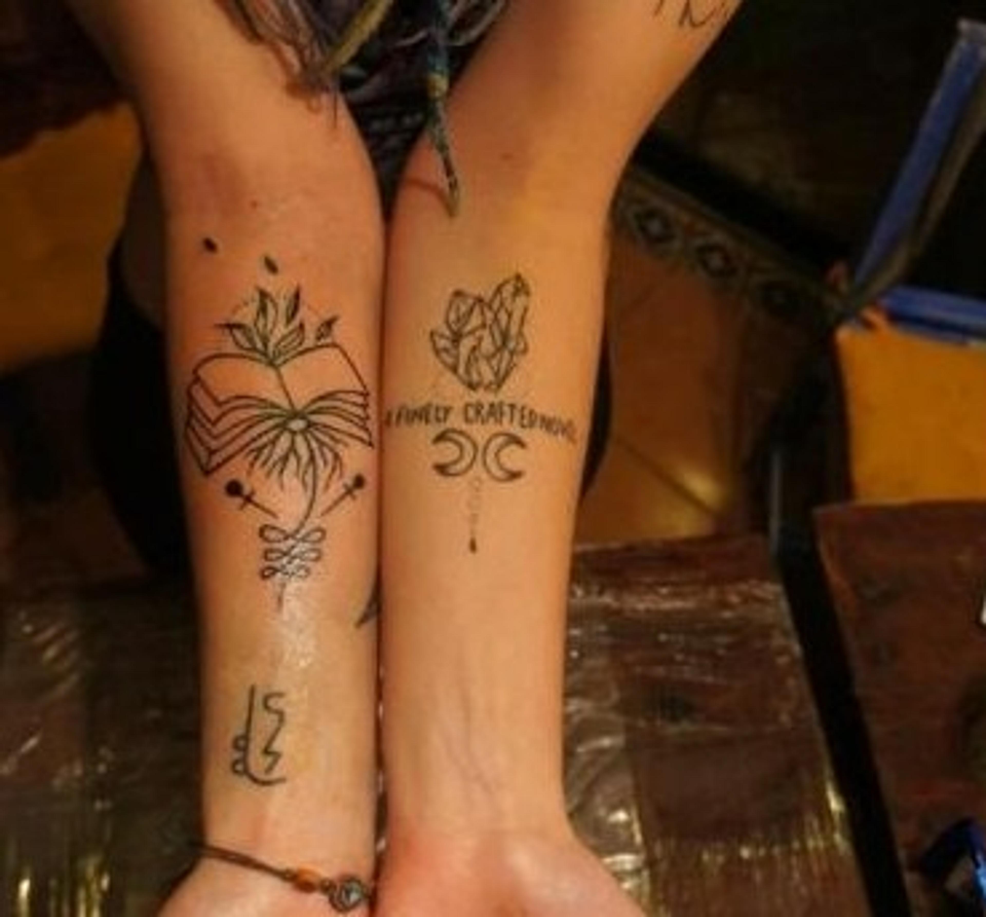 Intricate tattoos featuring botanical and mystical designs adorn two forearms against a background hinting at an artistic setting.