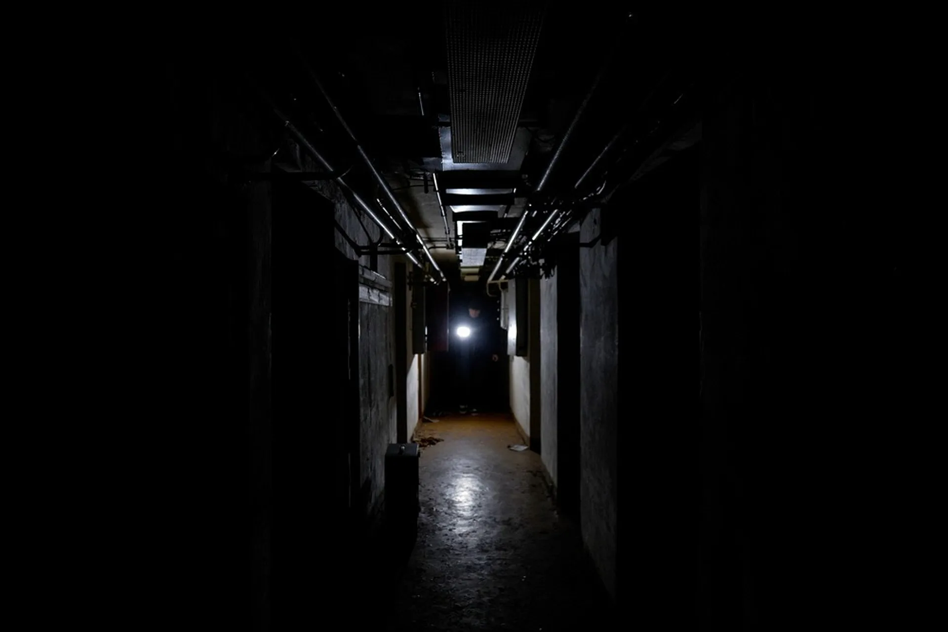 A person holding a flashlight stands at the end of a dark, narrow corridor.