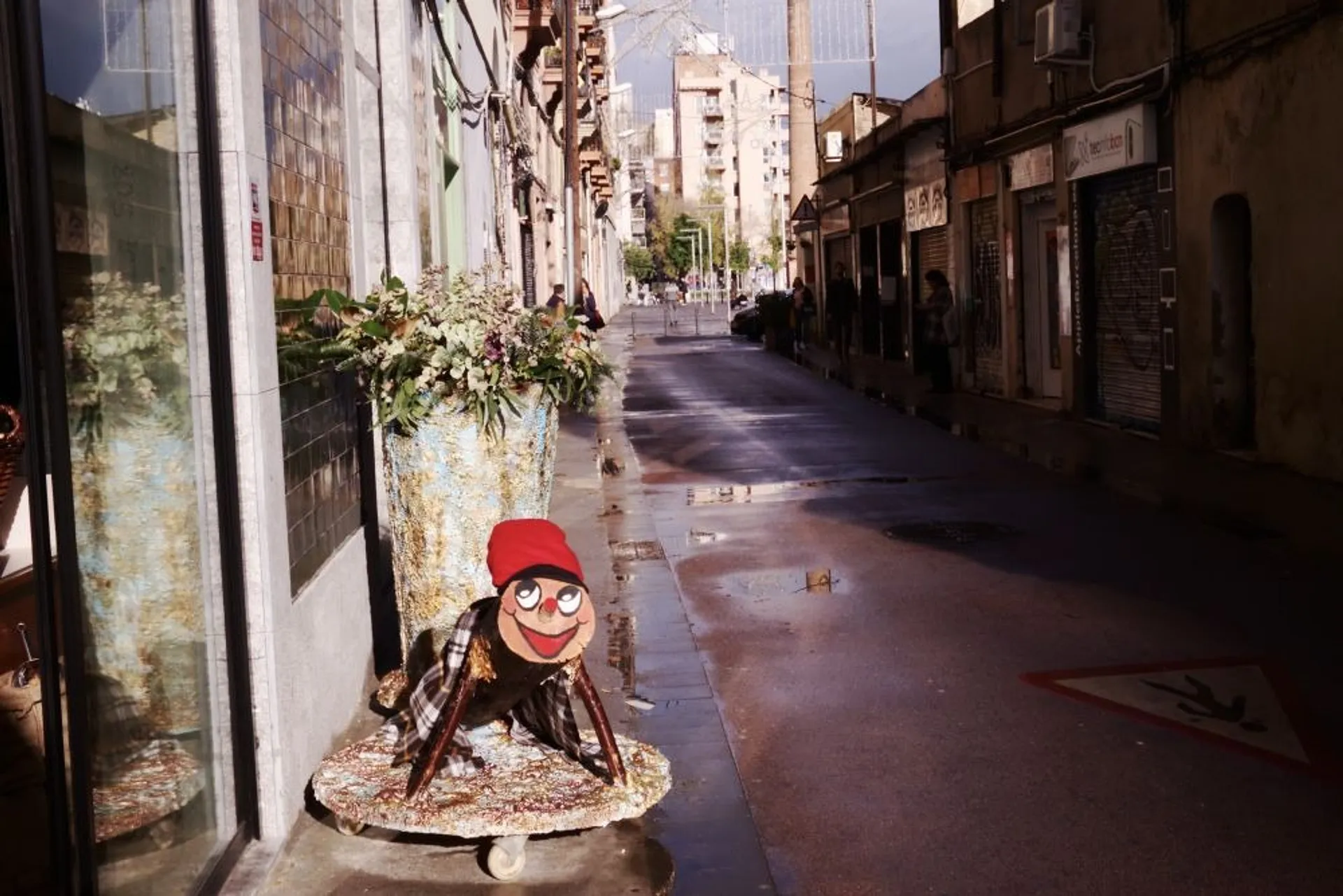 A narrow street in Barcelona features a whimsical wooden figure with a red hat beside a shopfront.